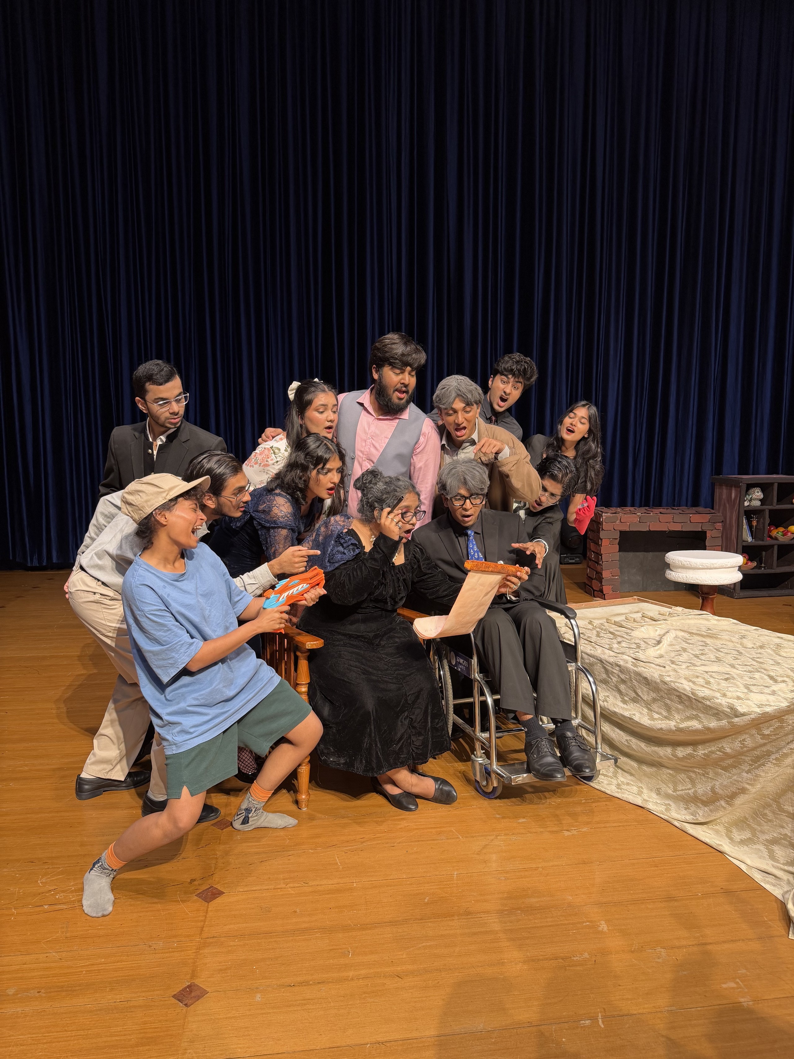 The full ensemble in rehearsal on stage: the family crowds around the wheelchair and the will, everyone singing.