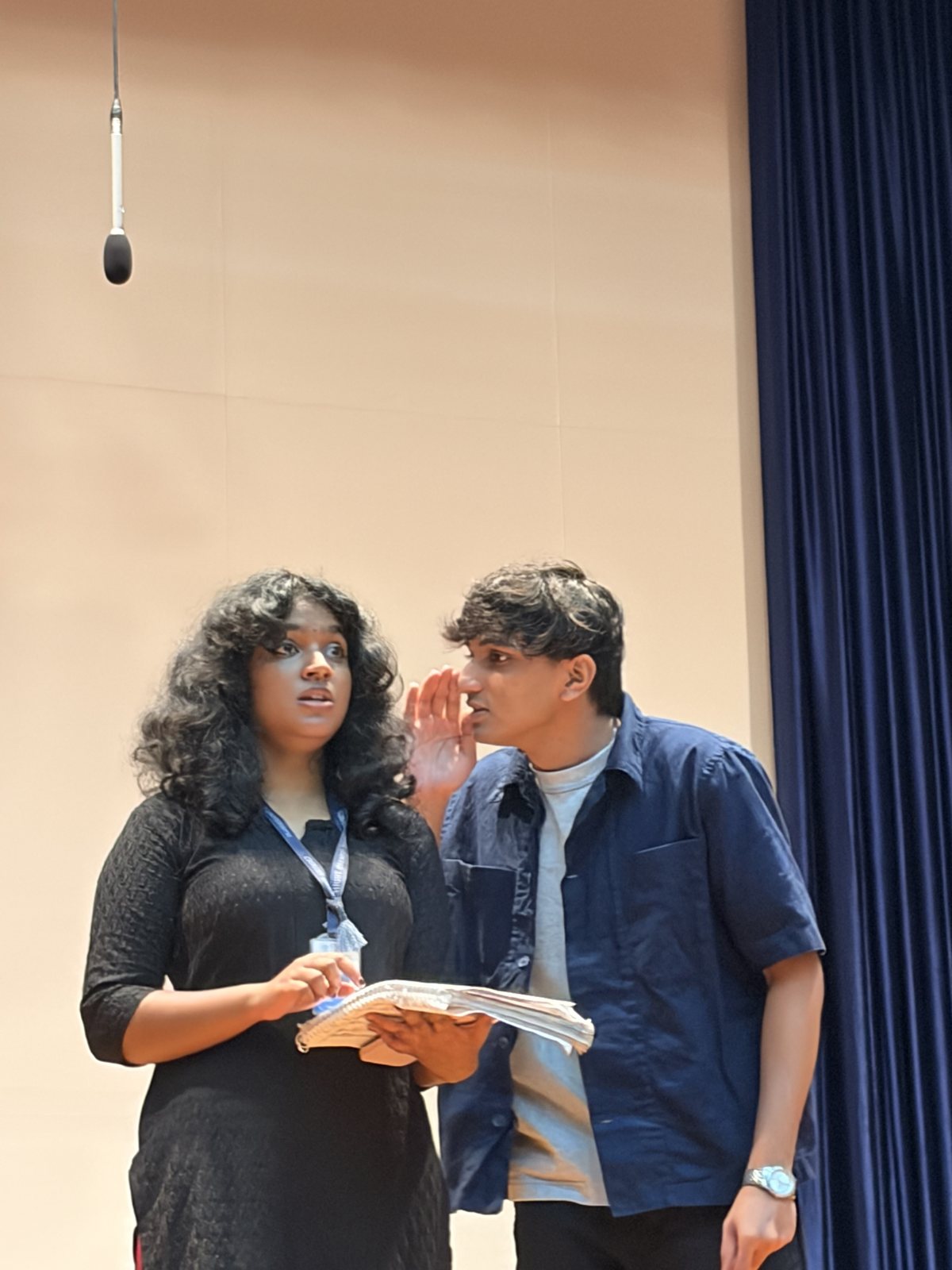 One singer leaning in to speak into another's ear, both holding their scores.