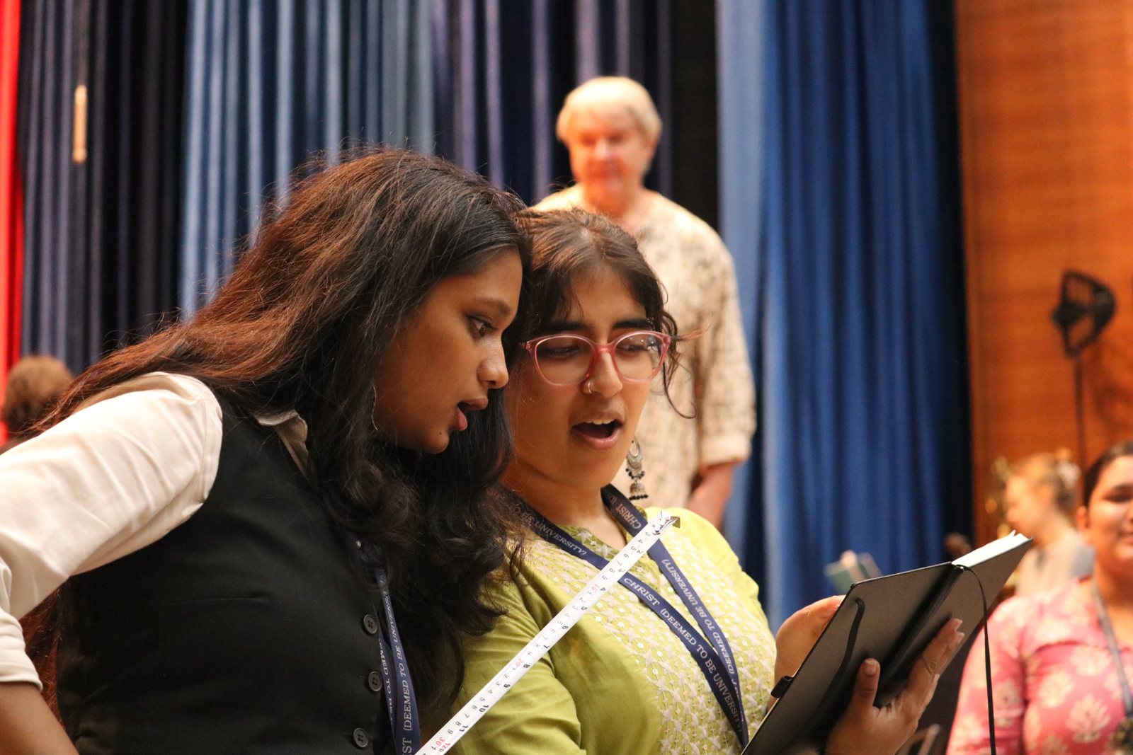 Two singers sharing one score mid-phrase, an older colleague watching from behind.