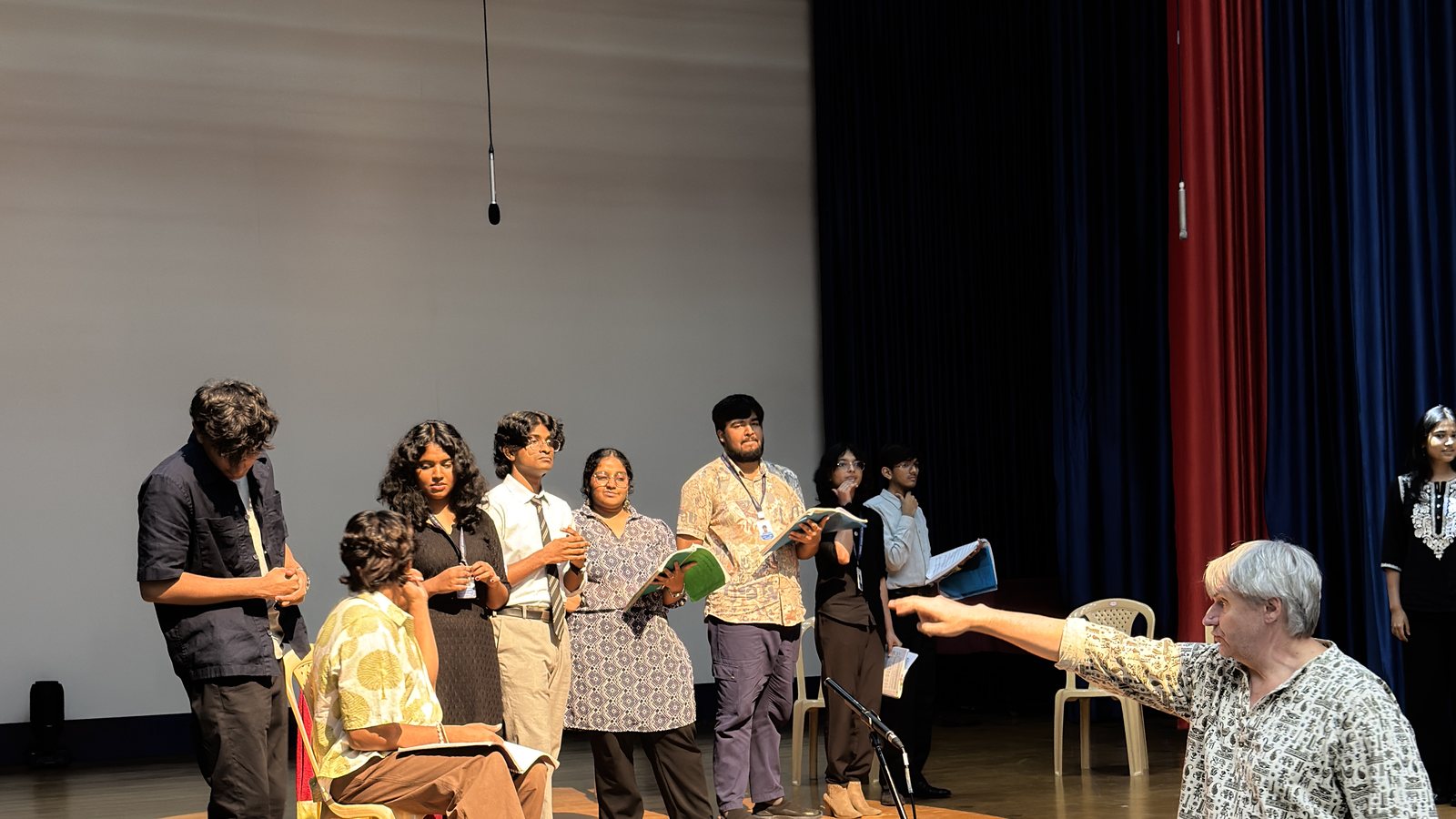 A row of the cast standing with their scores while a raised arm on the right gives a direction across the group.